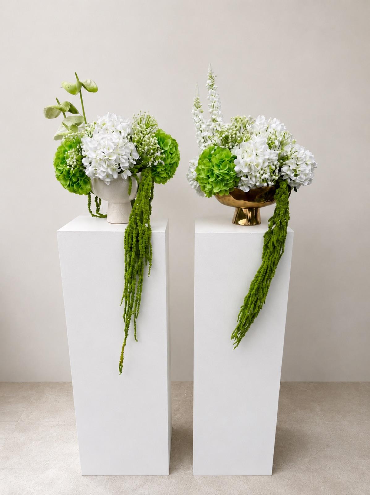 A lush faux floral centrepiece arrangement featuring white and green blooms with cascading trailing stems. Available in a white ceramic or gold bowl vase. Colours can be customized to suit your palette — perfect for wedding ceremony displays, reception tables, bridal showers, and elegant event styling. Pairs beautifully with our white display plinths.