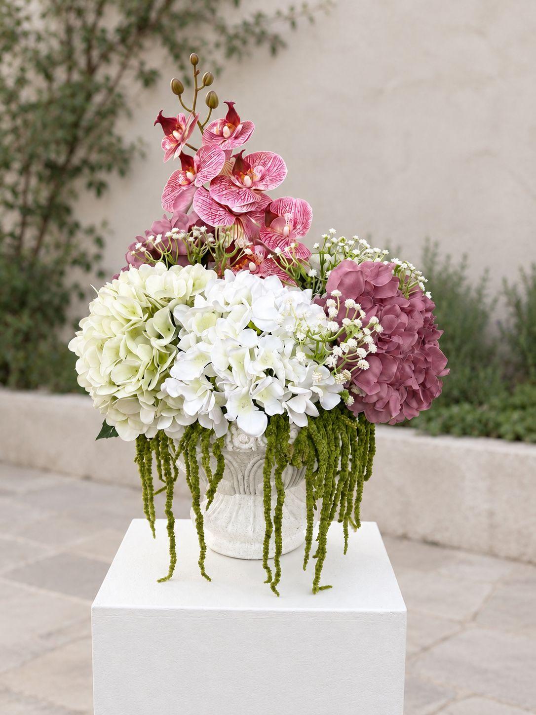 A romantic faux floral arrangement featuring mauve and ivory hydrangeas, striking pink orchids, delicate white accent blooms and cascading greenery in a textured white urn. Perfect for baby showers, ceremony entrances, welcome displays, signing tables, or anywhere you would like to add an elegant floral statement.